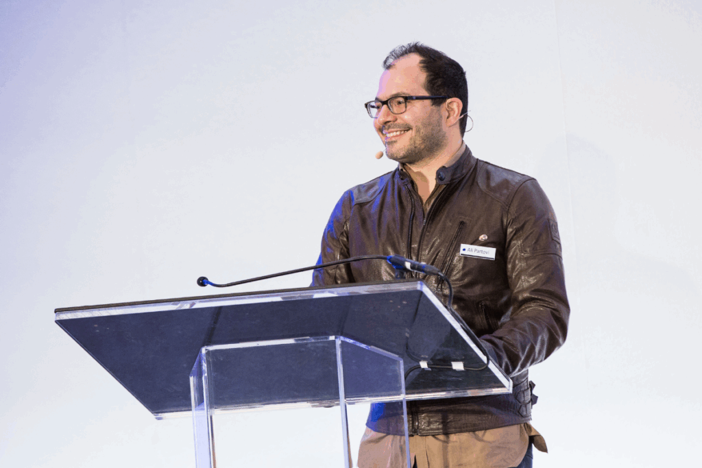 Ali Partovi presenting on stage, gesturing mid-speech while addressing a crowd of startup founders at a tech event.