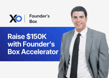 Smiling man in a gray suit standing beside a banner that says “Raise $150K with Founder’s Box Accelerator.”