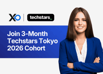 Smiling young woman in a blue blazer standing beside a banner that invites founders to join the 3-month Techstars Tokyo 2026 cohort.
