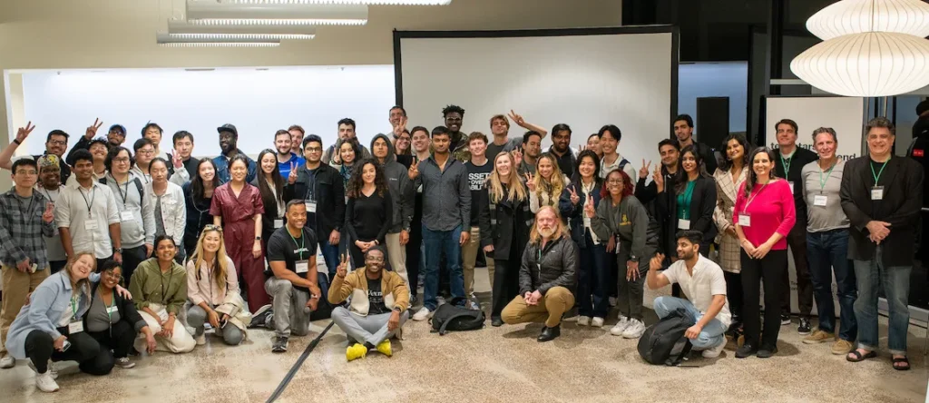 Large group of diverse founders, students, and mentors posing together at a startup event in front of a projection screen